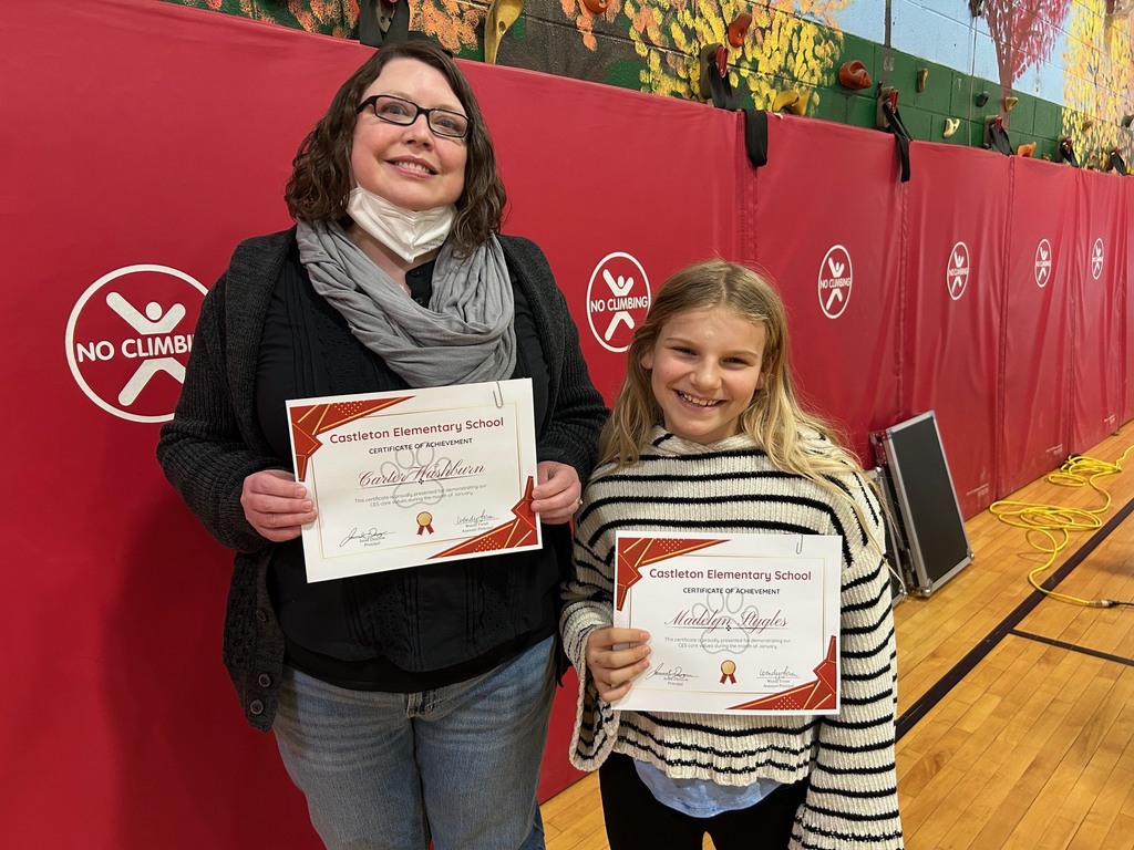 A staff member and a student stand together in front of red gym wall padding, each holding a Castleton Elementary School certificate of achievement and smiling.