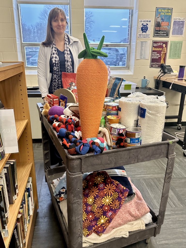 A teacher standing behind a rolling cart filled with donated pet supplies and items, including food cans, paper towels, toys, and a large carrot-shaped scratcher, collected for an animal shelter.