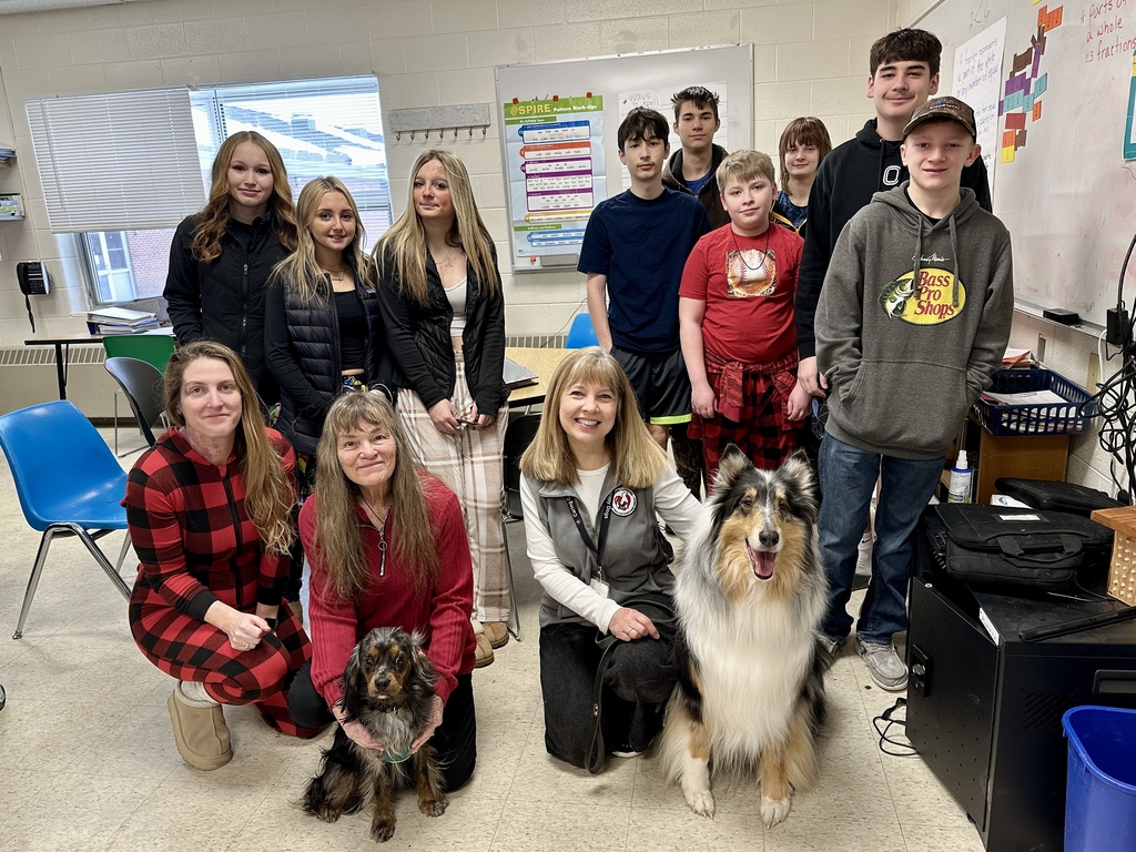 Group of middle school students and several adults in a classroom posing with two therapy dogs, one large collie-type dog and one small long-haired dog, during a school visit.