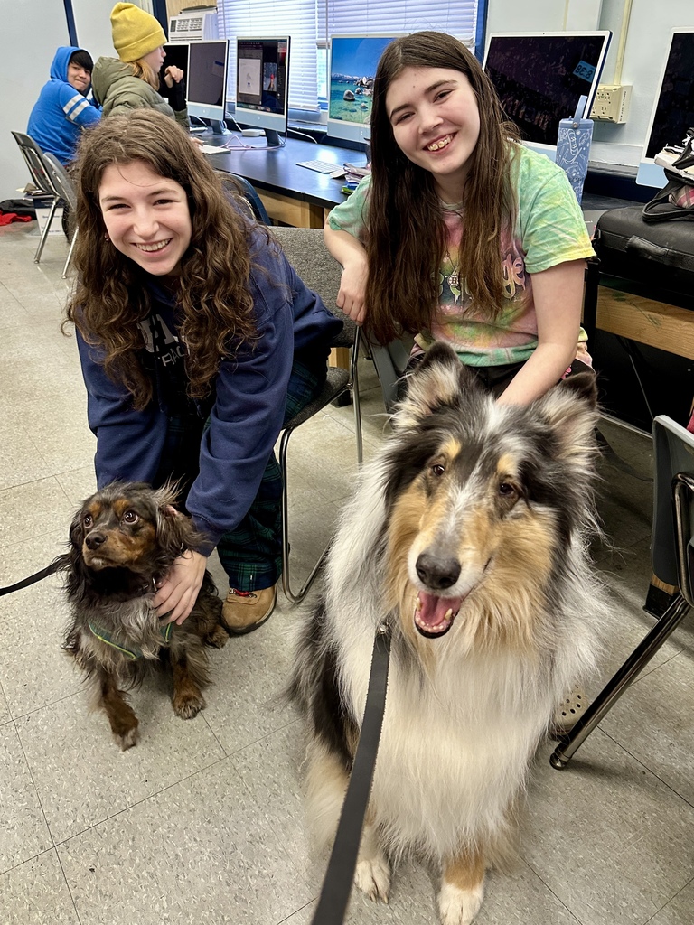 Two middle school students sitting in a computer lab smiling and petting two therapy dogs on leashes beside their chairs.