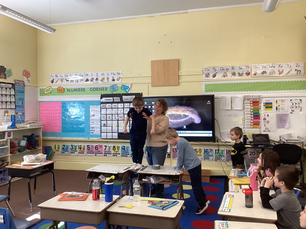 A teacher assists another student stepping onto desks to test a paper bridge, while classmates observe and classroom posters, number lines, and a word wall are visible behind them.
