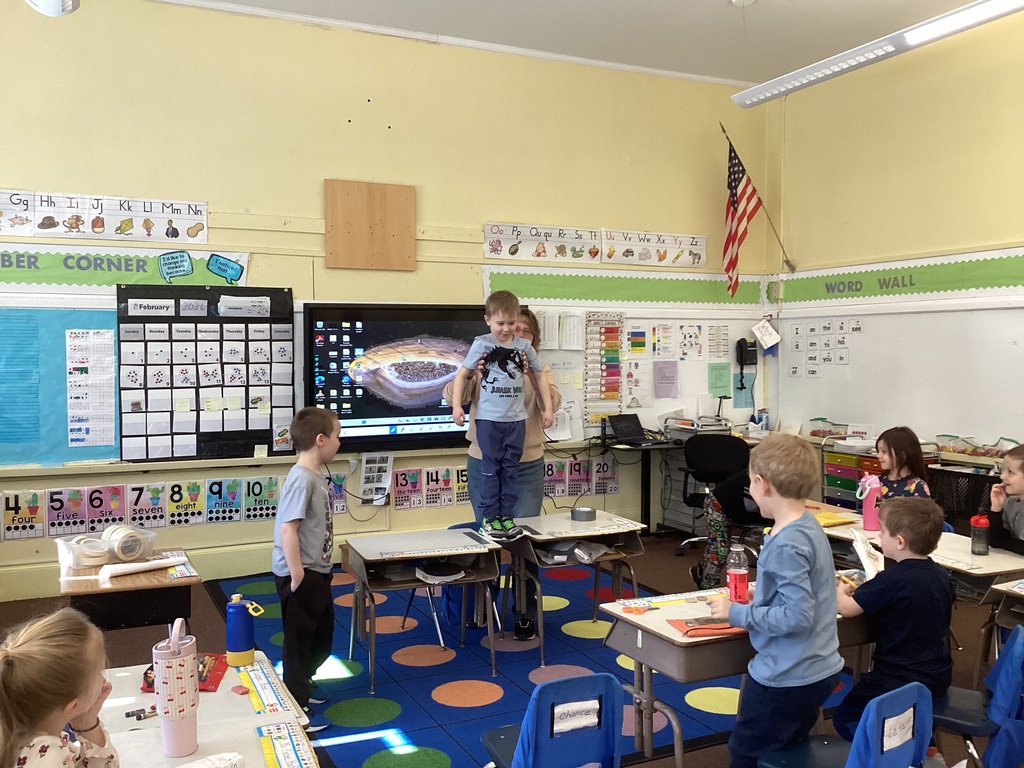 A teacher assists another student stepping onto desks to test a paper bridge, while classmates observe and classroom posters, number lines, and a word wall are visible behind them.