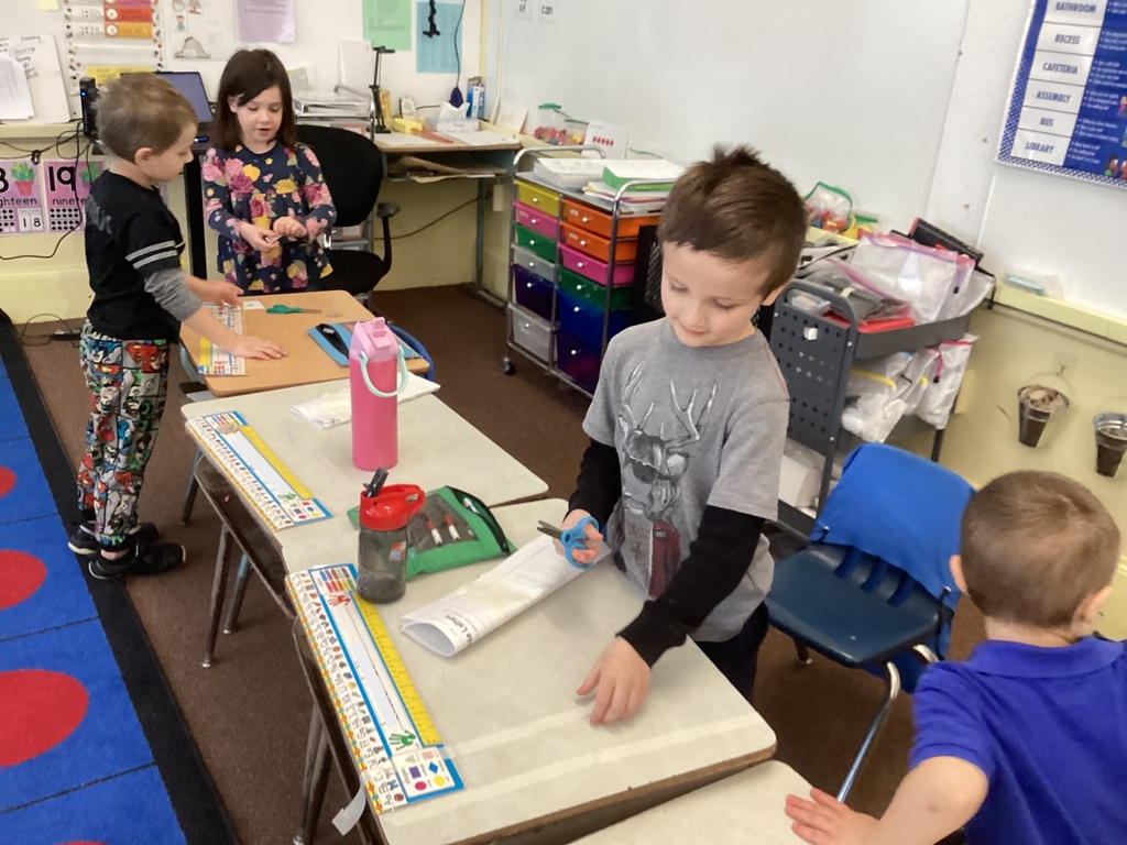Kindergarten students stand at their desks cutting and folding paper strips with scissors and tape while working on a classroom engineering challenge, with supplies and water bottles visible on the desks.