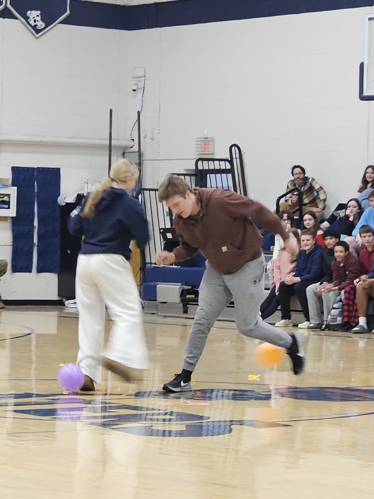 Two students race side by side across the gym floor, each guiding a balloon with their feet during a morning spirit challenge.