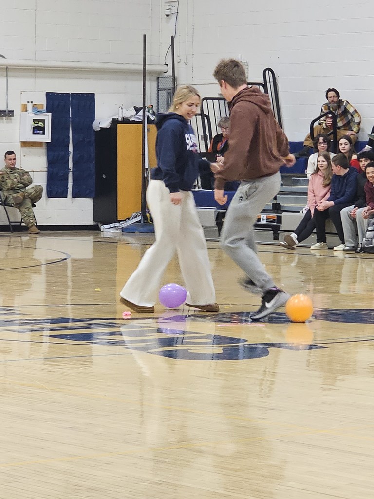 Two students laugh and maneuver around balloons at center court while other students sit in the bleachers behind them.