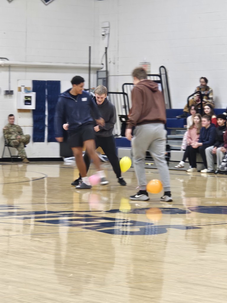 Three high school students in a gym compete in a spirit game, moving quickly while keeping balloons near their feet as classmates watch from the bleachers.