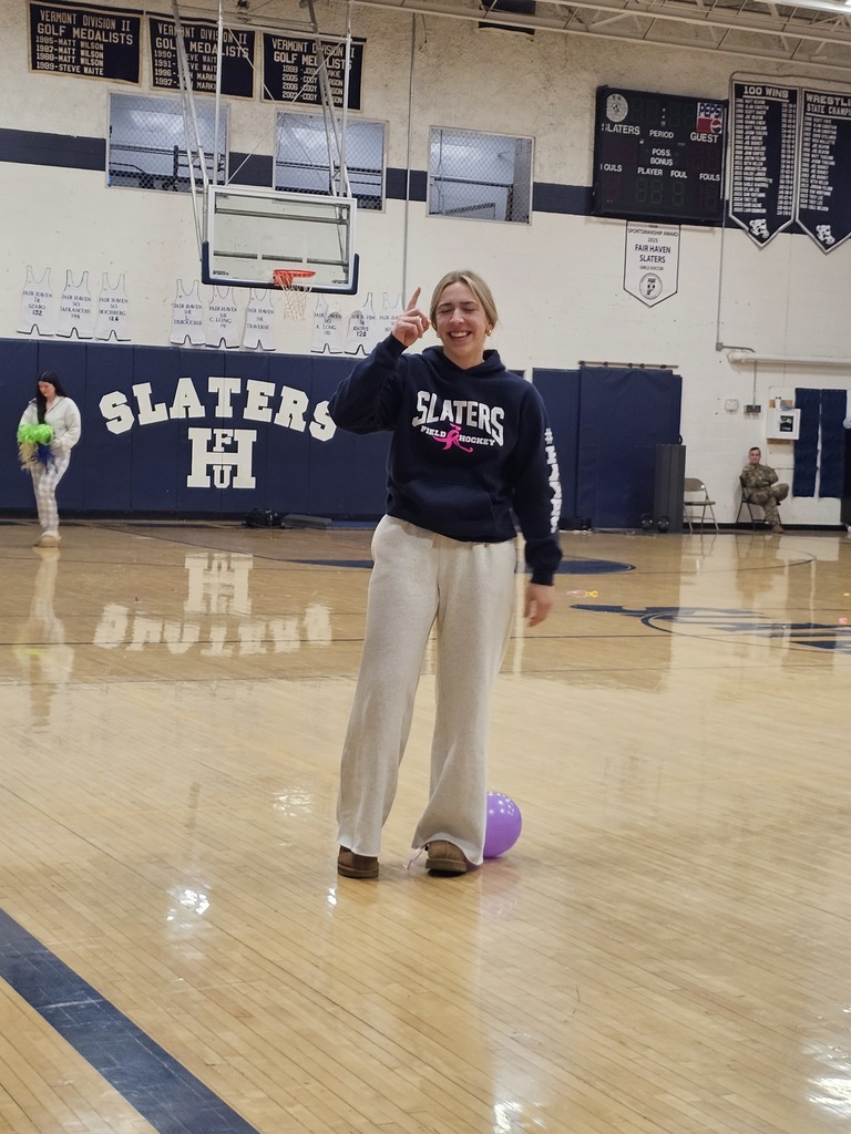 A student wearing a Slaters sweatshirt stands smiling on the gym floor with a balloon at her feet, celebrating after winning the spirit game.