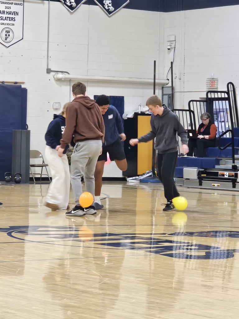 A small group of students on the gym floor step and pivot around balloons during a Winter Carnival spirit game while spectators look on.