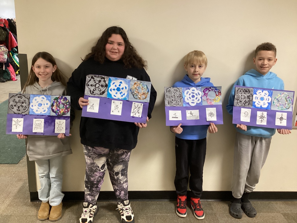Four elementary students pose in a hallway holding snowflake art project boards with mixed-media circular designs and small labeled sketches underneath.