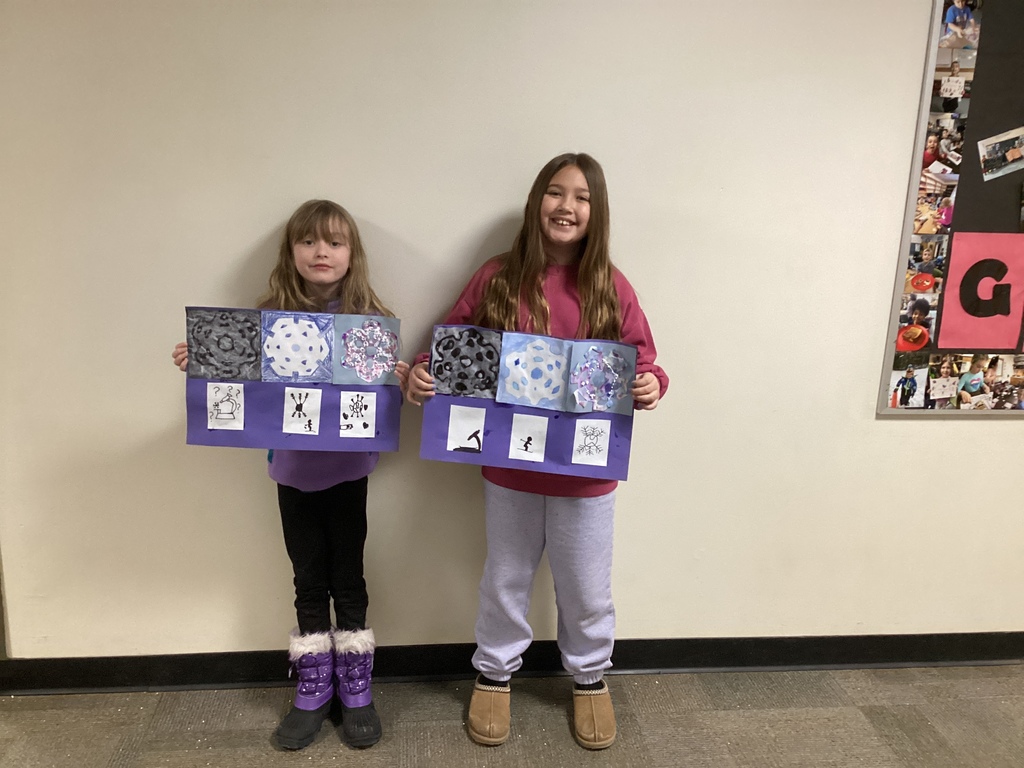 Two elementary students stand against a wall holding purple-backed snowflake art projects showing patterned designs and small sketch tiles below each finished piece.