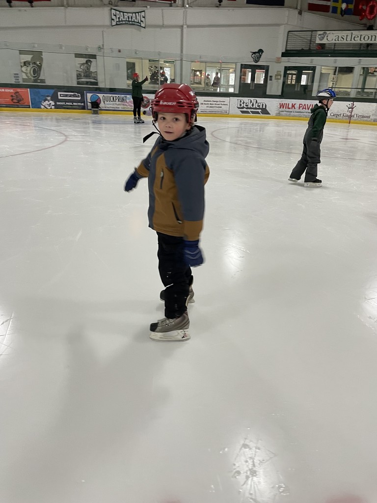 A young boy in a red hockey helmet and a brown and blue jacket standing on ice skates at an indoor rink. He is looking back over his shoulder toward the camera with a slight smile.