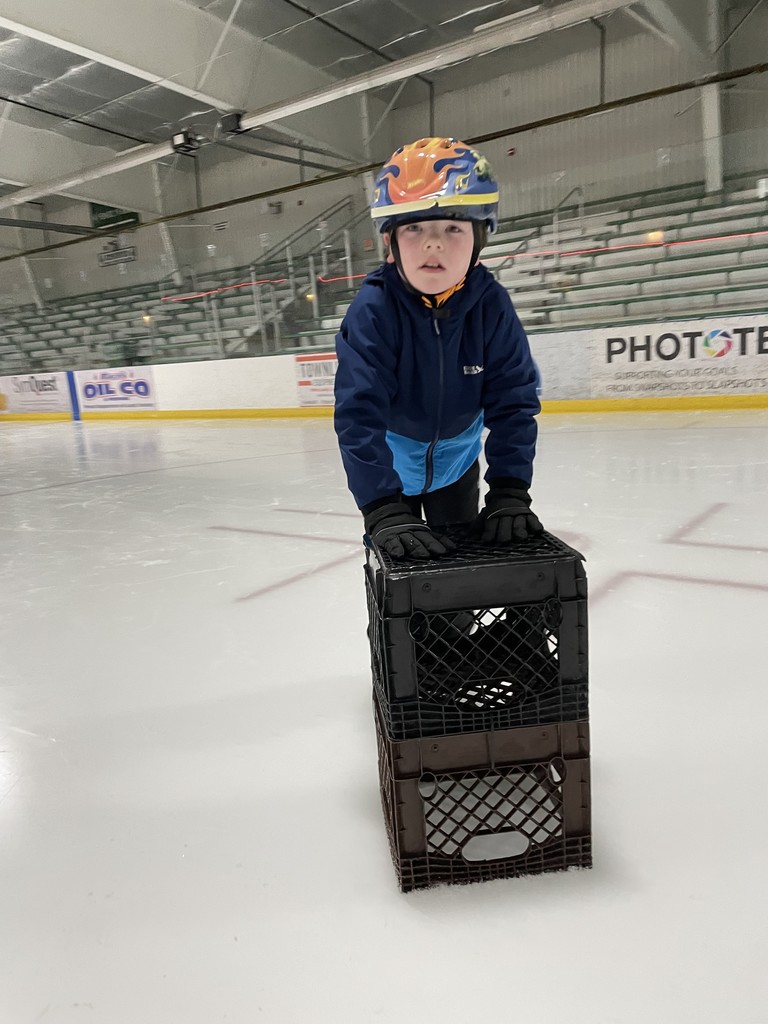 	A young boy wearing an orange and blue flame-patterned helmet uses two stacked plastic milk crates as a stabilizer to help him balance while ice skating.