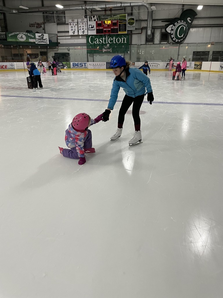 An older girl in a blue jacket and blue helmet reaches down to hold the hand of a toddler in a pink helmet and purple snowsuit who has fallen on the ice. A scoreboard in the background shows the "Castleton" rink name. 