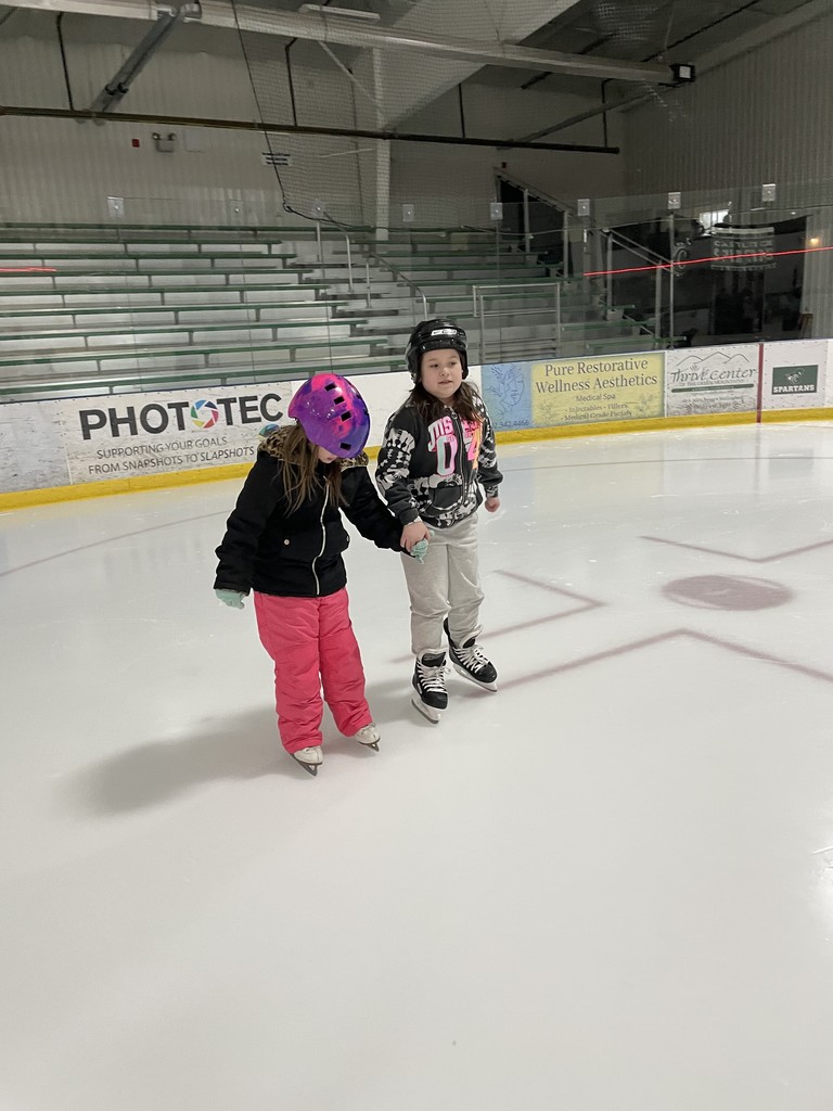 	Two young girls skating together on an indoor rink. One wears a vibrant pink and purple helmet and pink snow pants; the other wears a black helmet and grey pants. They are holding hands for balance. 