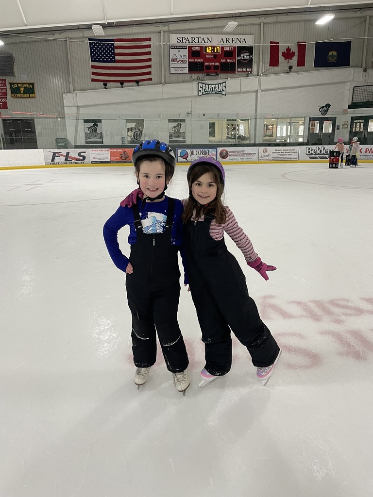  IMG_5465.jpg Two young girls in ice skates and helmets pose together on an ice rink. The girl on the left wears a blue helmet, a blue long-sleeve shirt, and black snow bibs. The girl on the right wears a purple helmet and a pink striped shirt with black snow bibs. In the background, flags and a scoreboard for "Spartan Arena" are visible.
