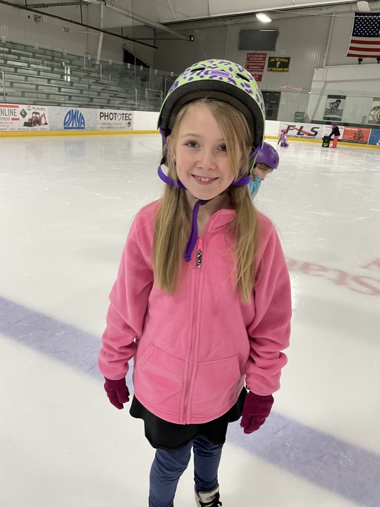 A close-up of a young girl with long blonde hair smiling on the ice. She is wearing a bright pink fleece zip-up jacket, a black skirt over blue leggings, and a green and purple leopard-print helmet.  