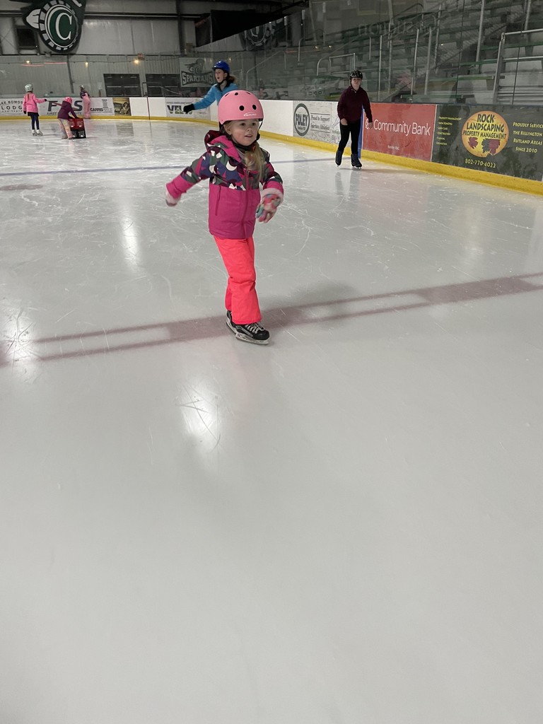 A young girl in a pink helmet, a colorful patterned winter jacket, and bright pink snow pants ice skates across the rink. She is captured in motion with her arms slightly out for balance. Other skaters and rink advertisements are visible in the background.  