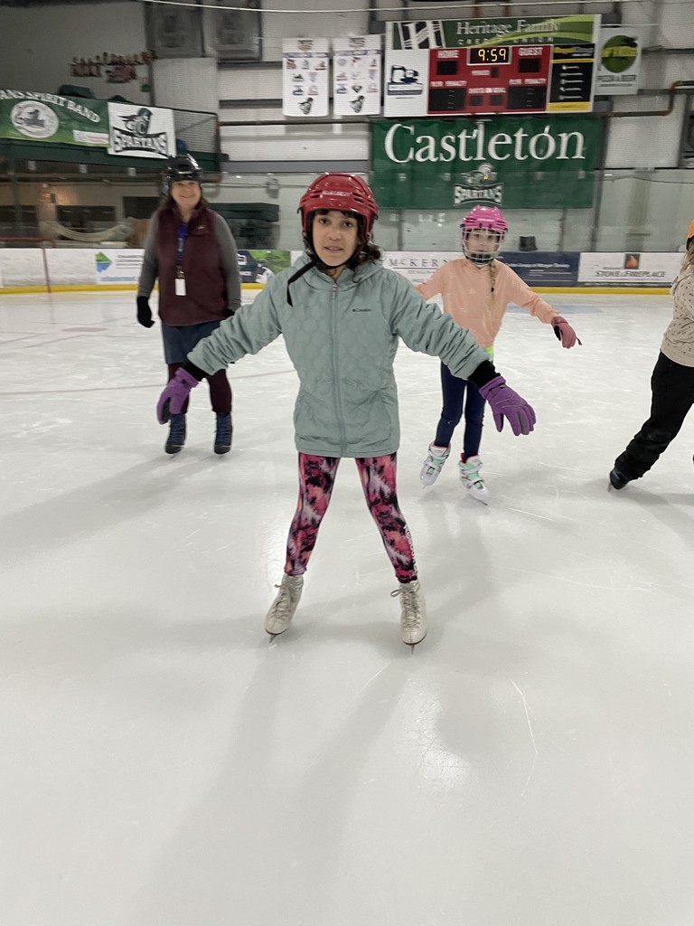 A young girl in a red hockey helmet and a light teal quilted jacket skates toward the camera with her arms outstretched for balance. She is wearing vibrant pink and black patterned leggings. Behind her, other children and an adult are skating under a large "Castleton" banner.  