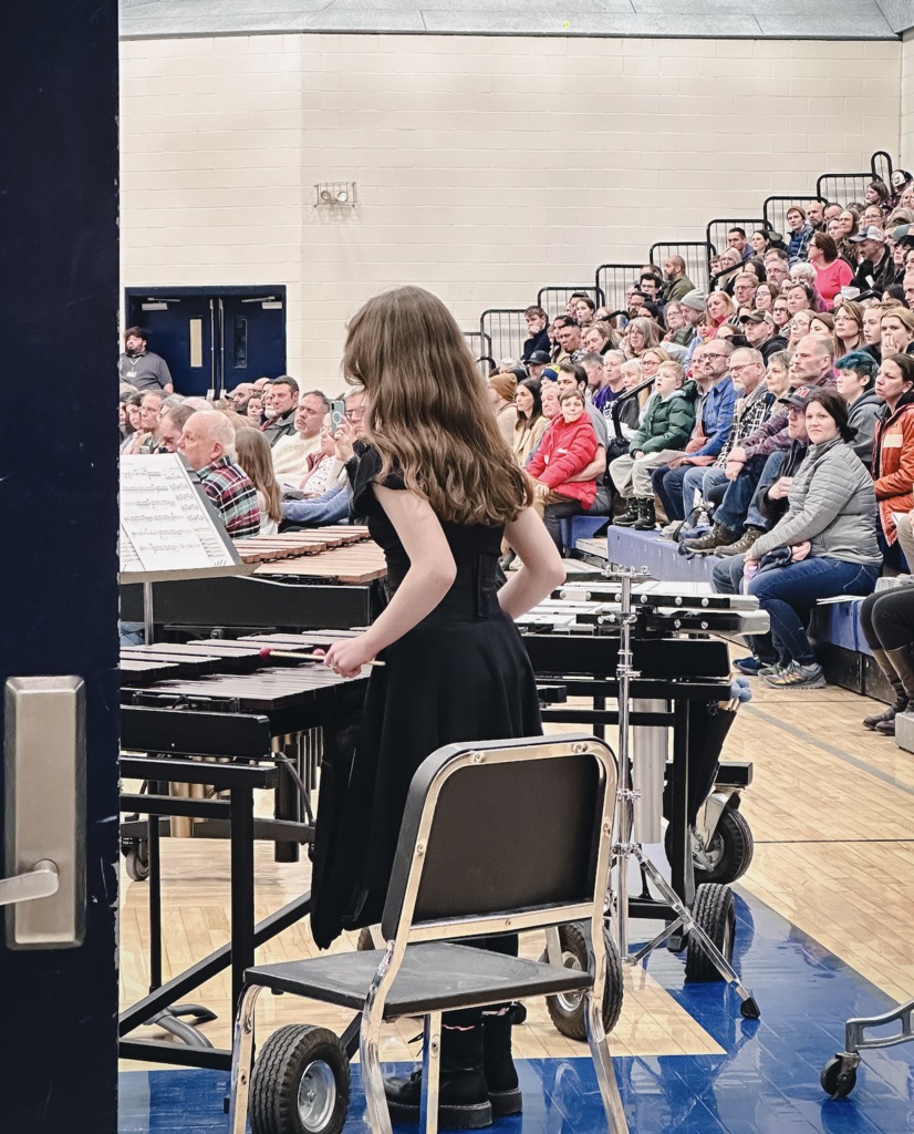 Student percussionist in black concert attire plays a mallet instrument near the gym floor while an audience watches from the bleachers behind.