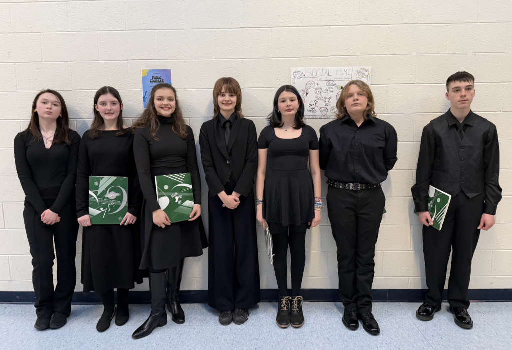 Seven middle school chorus students stand in a row against a hallway wall wearing black concert attire and holding green music folders, posing before a performance.