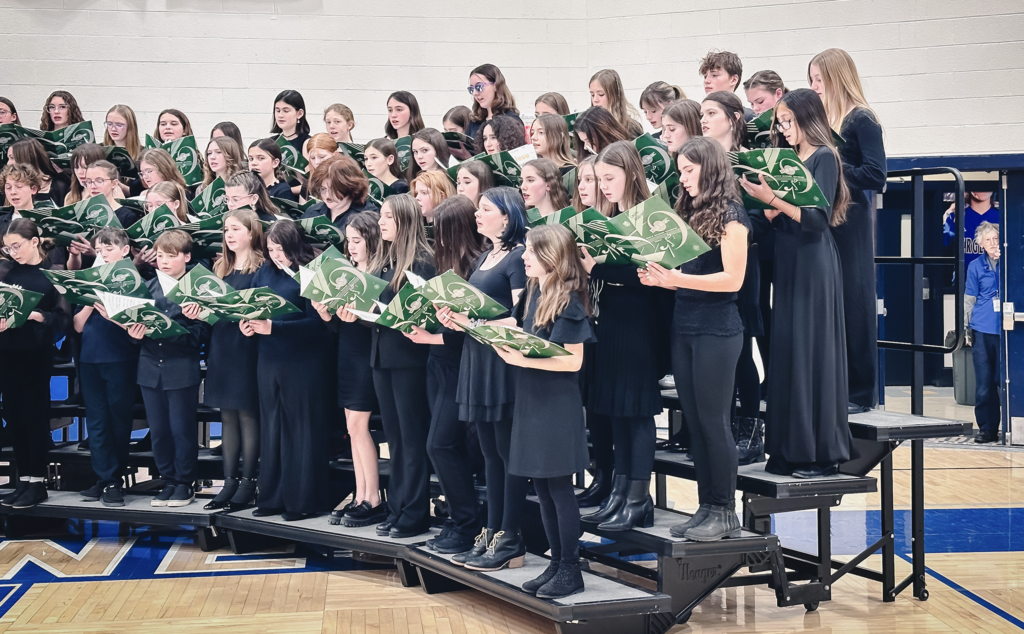 Close-up of a section of the student chorus on risers, dressed in black and singing while reading from green music folders.