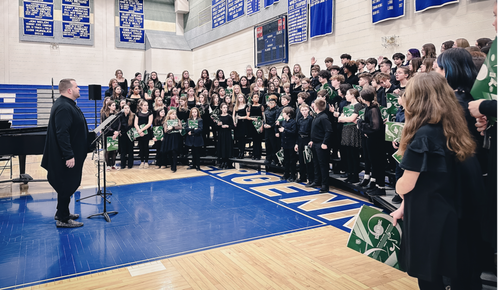 A large student chorus stands on risers in a school gym while a conductor faces them at a microphone stand; students wear black concert clothing and hold green folders.
