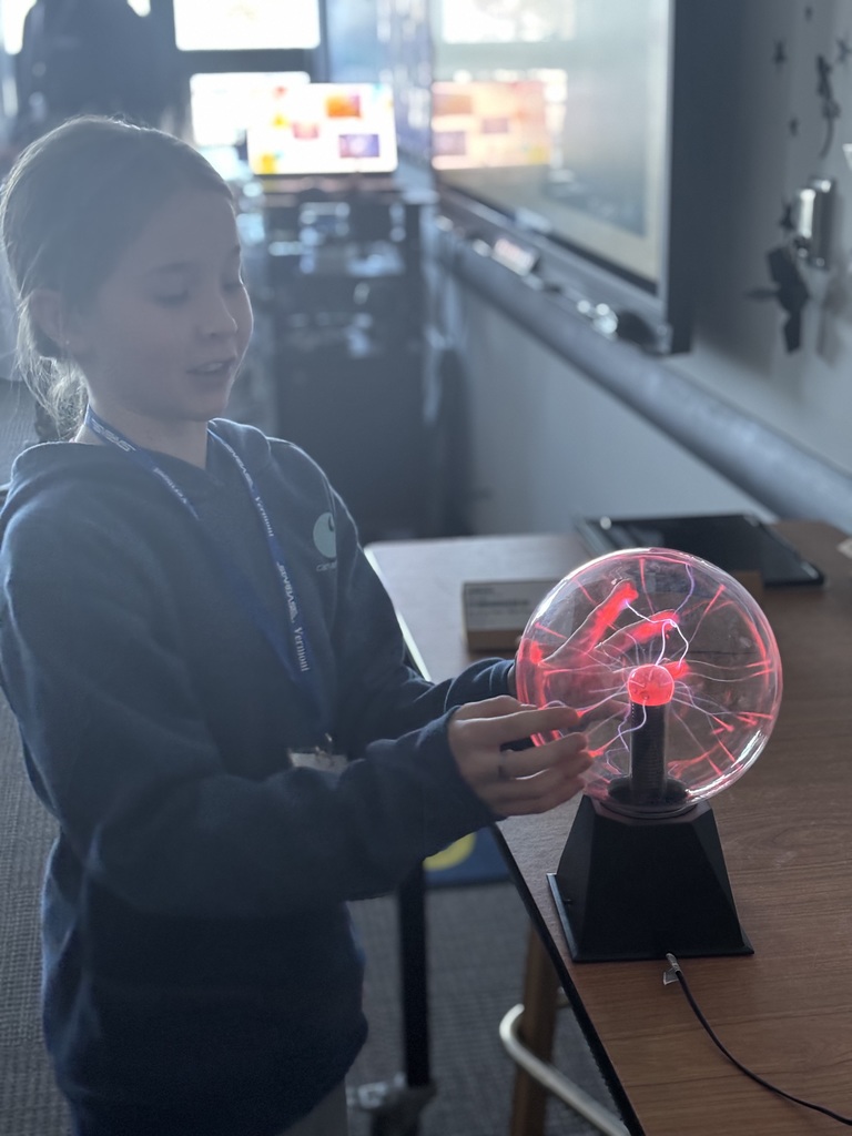 Student touching a glowing plasma globe on a table in a dim classroom while observing the electric light patterns inside the sphere.