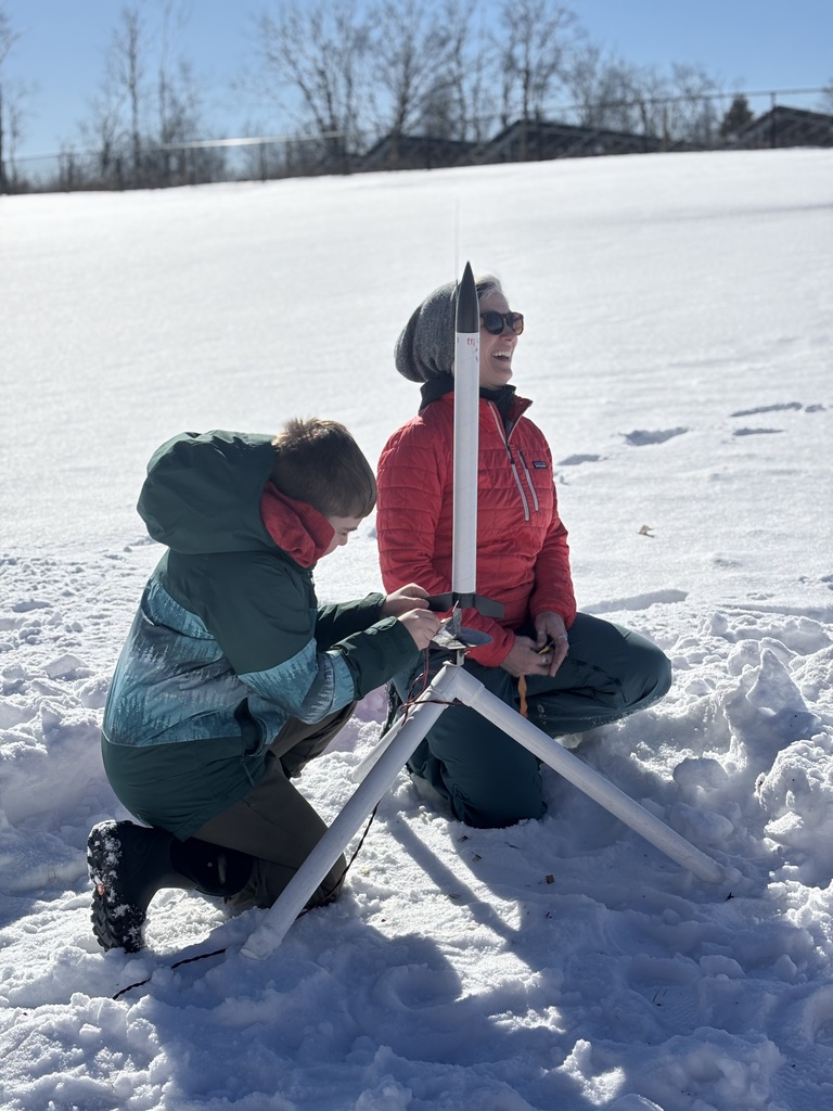 Student and adult kneeling in the snow outdoors setting up a model rocket on a launch stand under a bright blue sky.