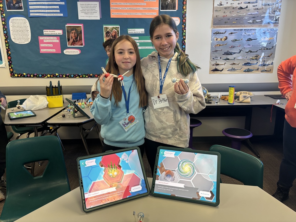 Two students standing together in a classroom holding molecular models, with two tablets on a table showing oxygen and hydrogen science visuals.
