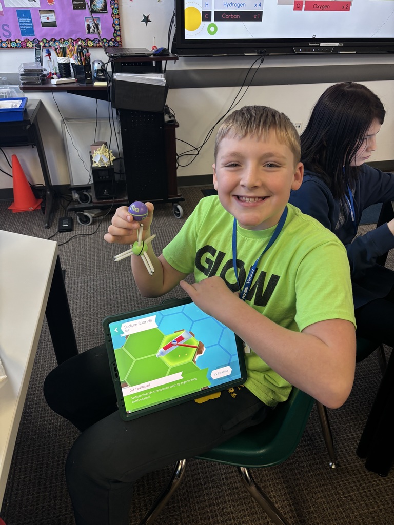 Smiling student seated in a classroom holding a molecular model while a tablet displays a sodium fluoride science graphic.