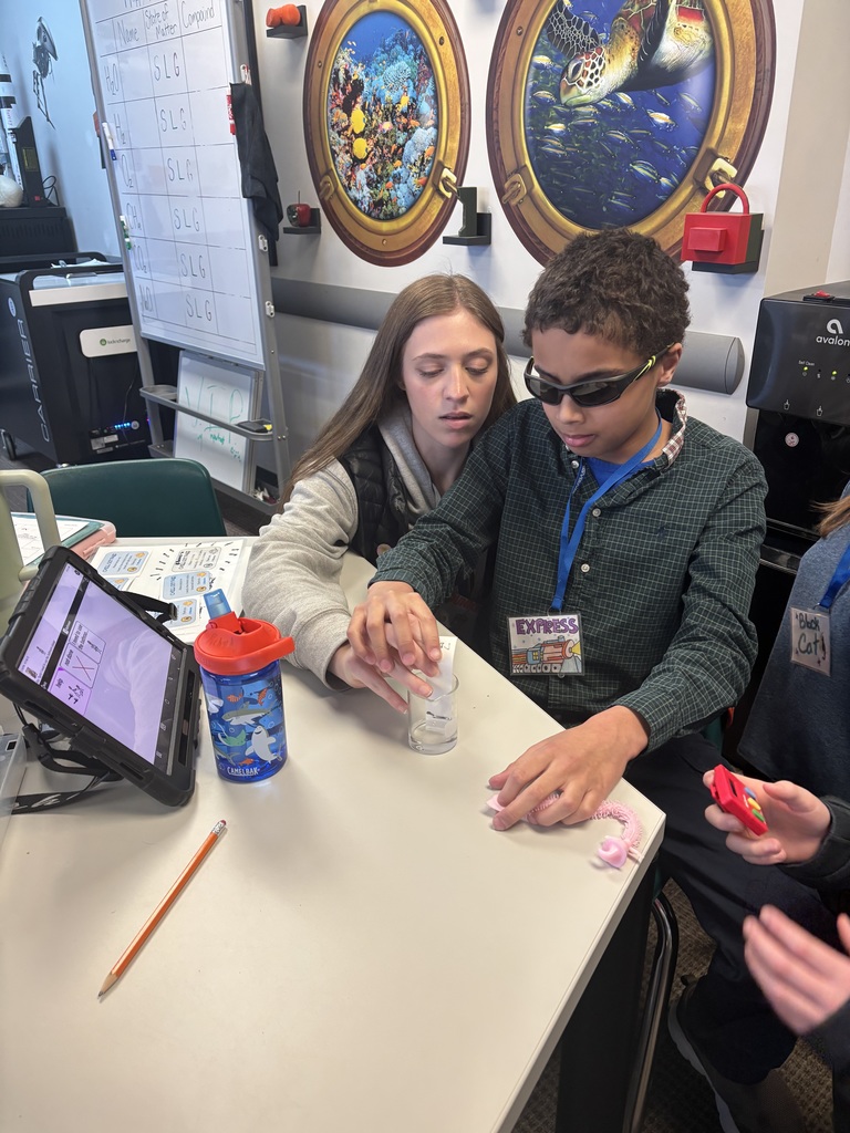 An adult and student at a table conducting a small experiment with a clear cup and tablet, while another student holds a device nearby.