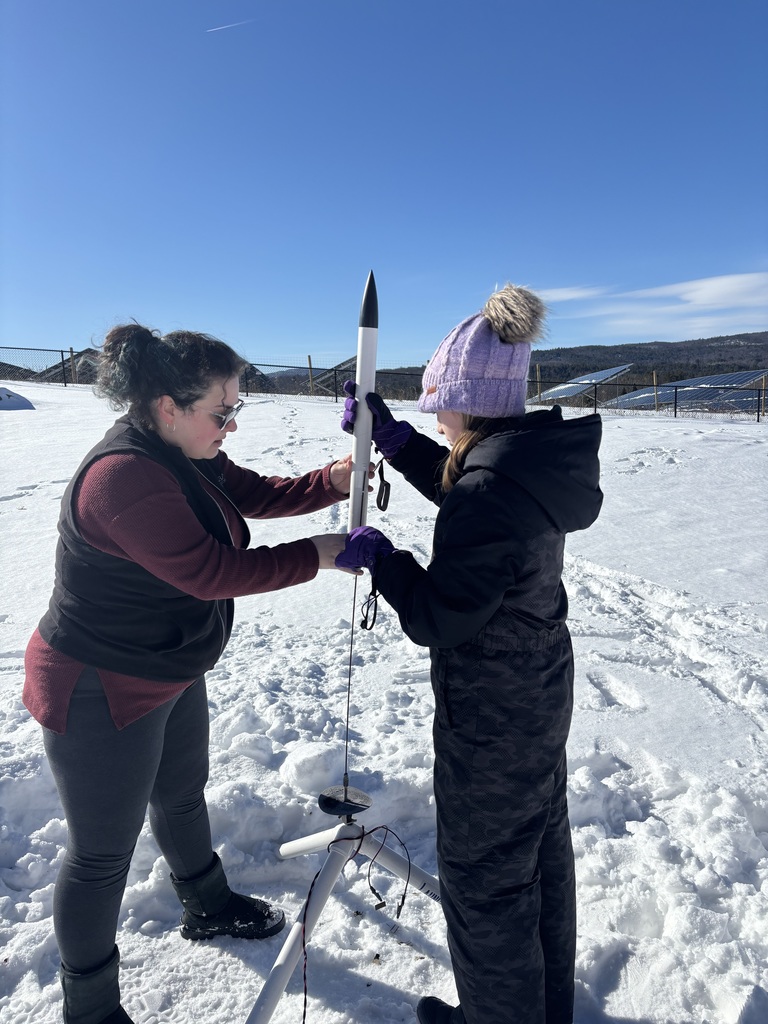 Adult and student in winter clothing preparing a model rocket for launch in a snowy outdoor area near a fence and solar panels.