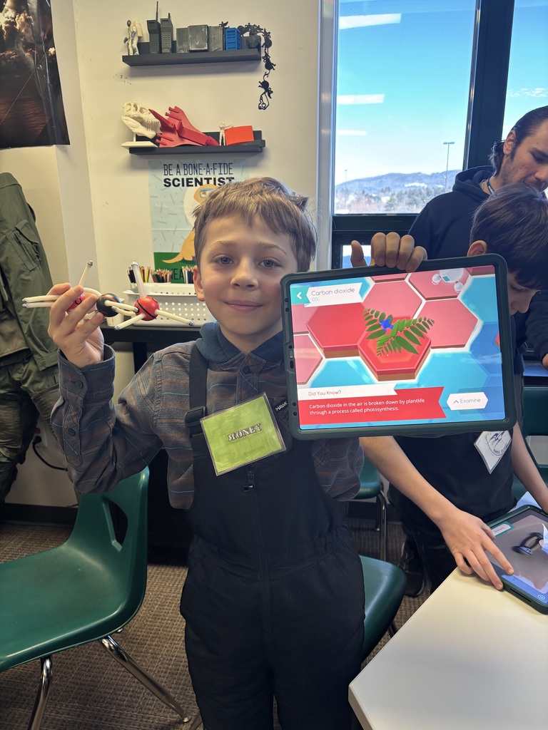 Student in a classroom holding a molecular model and a tablet showing a carbon dioxide and photosynthesis diagram, with science posters and shelves in the background.