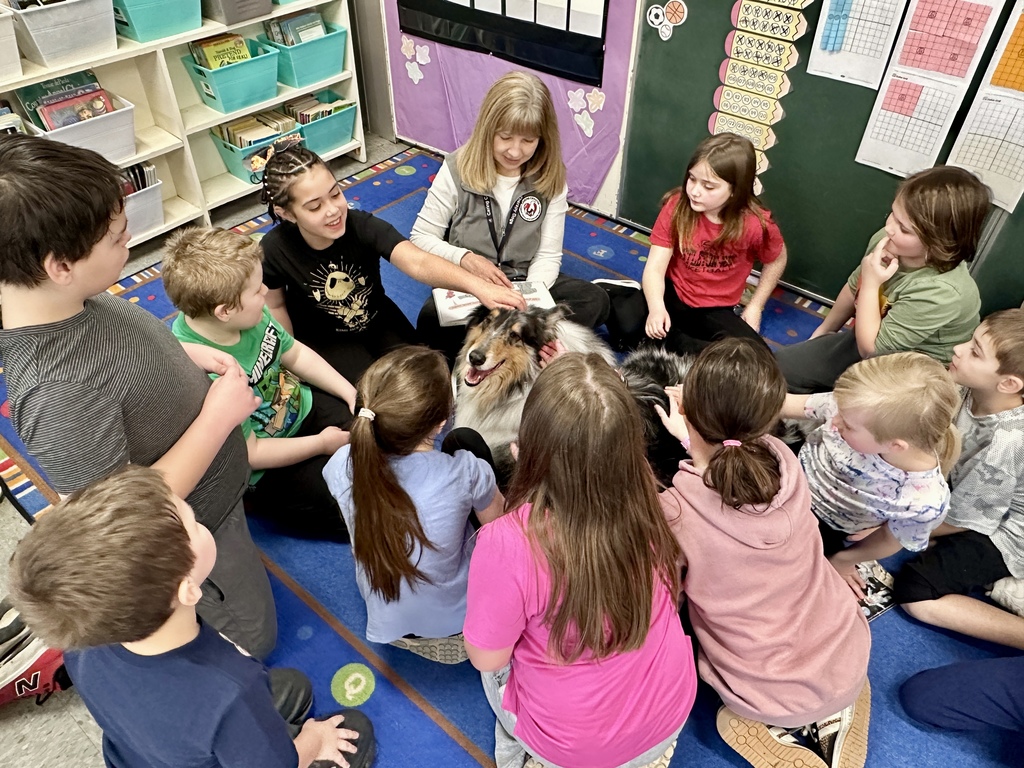 Classroom circle of students sitting around a smiling collie therapy dog while an adult handler reads and guides the activity.