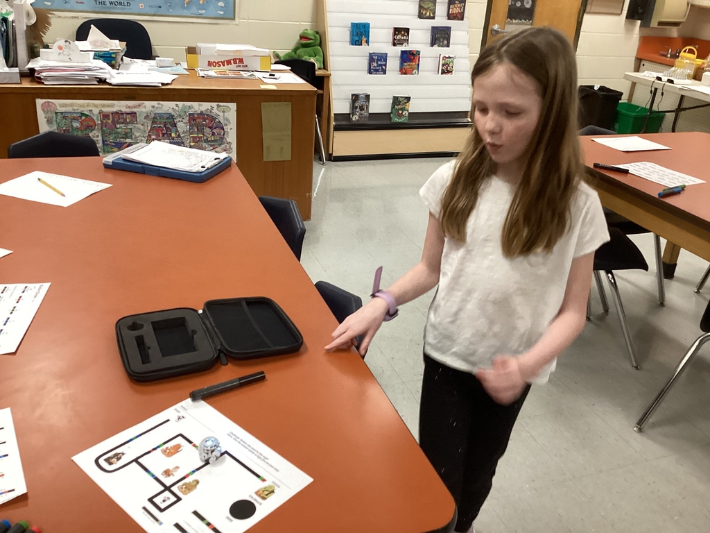 A student stands at a table guiding a small Ozobot robot along a color-coded path on a worksheet, with markers and a robot case nearby in the library classroom.