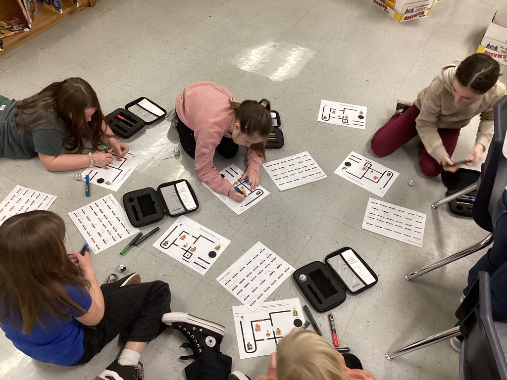 A group of students spread out on the floor in a circle, drawing colored coding lines on printed maps and programming sheets, with Ozobot cases and markers placed around them.