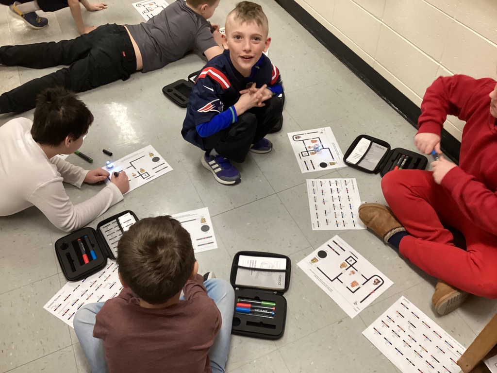 Several fourth grade students sit and kneel on the classroom floor working with papers and Ozobot coding sheets, using colored markers to draw code paths while a small robot sits on the page.