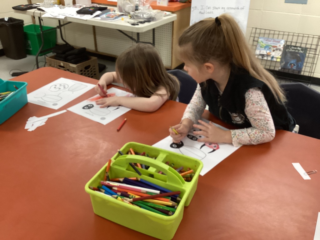 Two Pre-K students sit side by side at a table coloring beetle puppet templates, with a green caddy of colored pencils in the foreground.