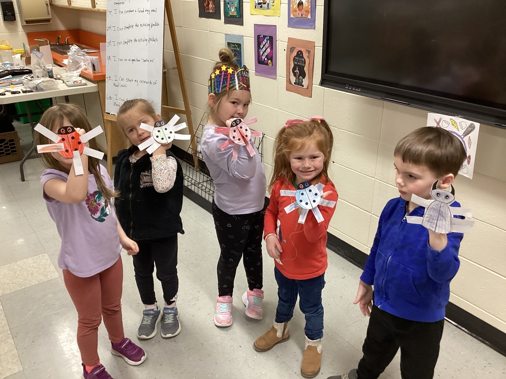Five Pre-K students smile and display their decorated beetle puppets on sticks while standing together in the library classroom.