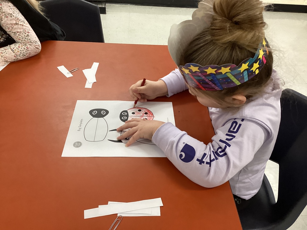 A student wearing a star headband colors a beetle puppet worksheet with a red crayon at a classroom table.