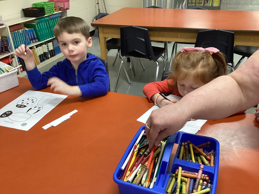 Two young students sit at a library table coloring beetle worksheets with crayons and colored pencils while an adult hand reaches into a supply box.