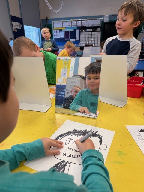 A child draws a detailed self-portrait while smiling at their reflection in a tabletop mirror; classmates work in the background.