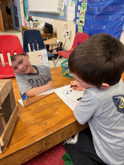 A child smiles at their reflection in a small mirror while sketching a face on paper at a wooden table.