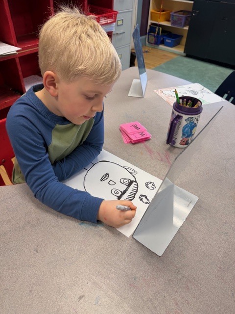 A student concentrates on drawing a face worksheet while looking into a small mirror on a desk, with crayons and supplies nearby.