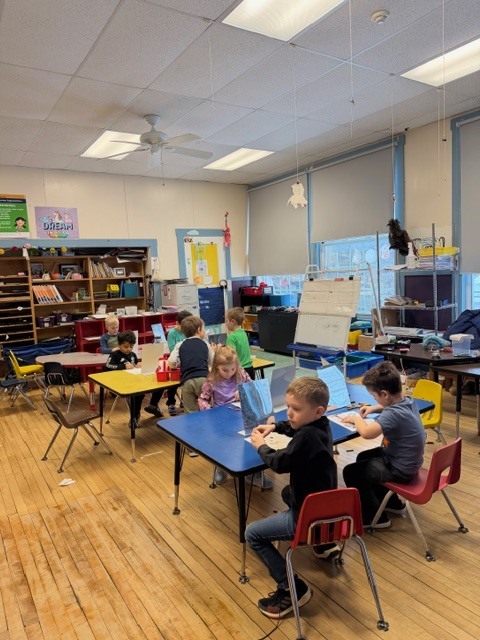 Wide classroom view of multiple students seated at different tables, each using a mirror to create self-portrait drawings with art supplies.