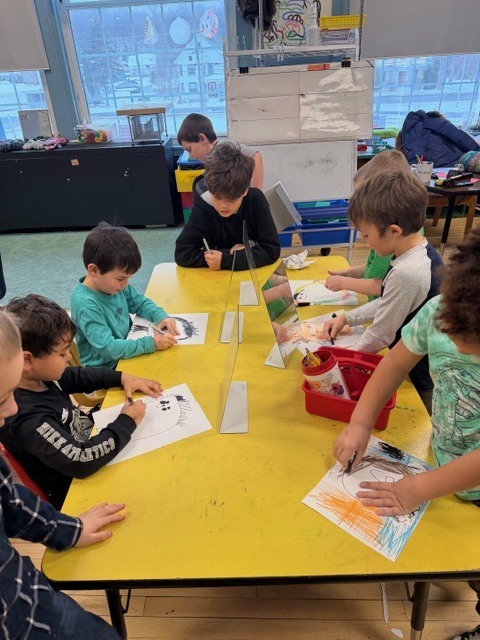 A group of young students at a yellow table color self-portrait worksheets while looking into upright mirrors.