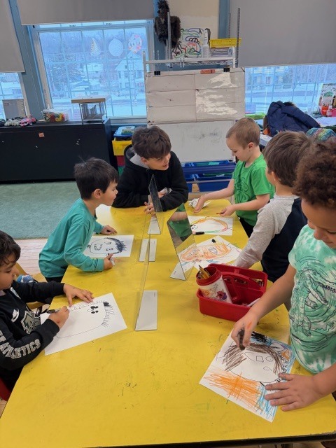 Several children compare their drawings to their reflections in mirrors while working together on self-portrait art at a shared table.