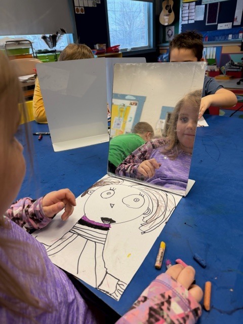 A student in a purple shirt studies their reflection in a mirror while coloring a self-portrait with crayons at a blue table.
