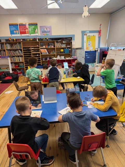 Elementary students sit at colorful tables in a classroom, using small tabletop mirrors to draw self-portraits with markers and crayons.