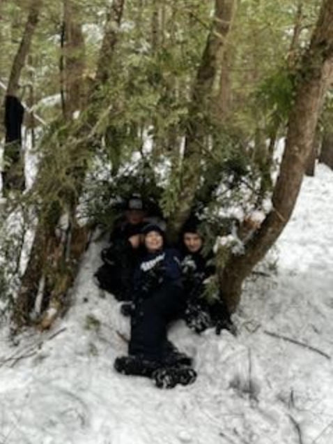 Three students sit inside a finished branch-and-bough shelter in the snowy woods.